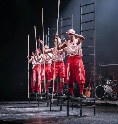 Een groep artiesten op krukken, die lange stokken vasthouden, gekleed in rood-witte kostuums op het podium.