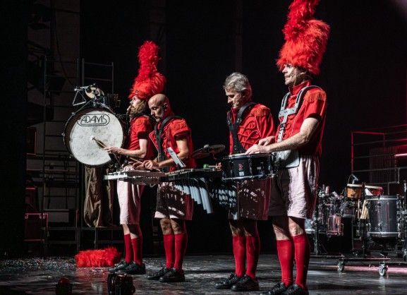 Vier muzikanten in rode uniformen bespelen percussie-instrumenten op het podium.