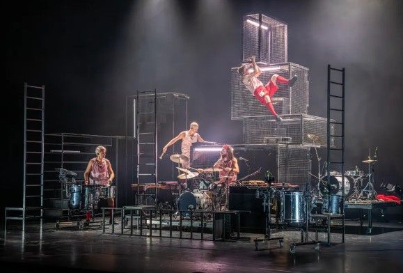 Muzikale artiesten op het podium met drumstel, metalen kooien, ladders en een acrobaat.