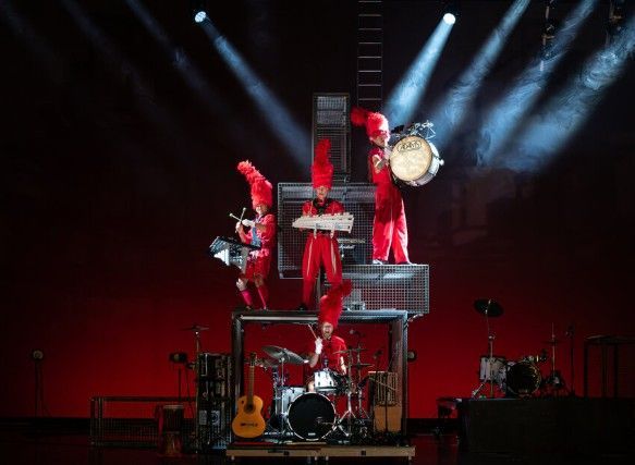 Muzikanten in rode uniformen op een podium met meerdere niveaus, drums en instrumenten, met een rode achtergrond.