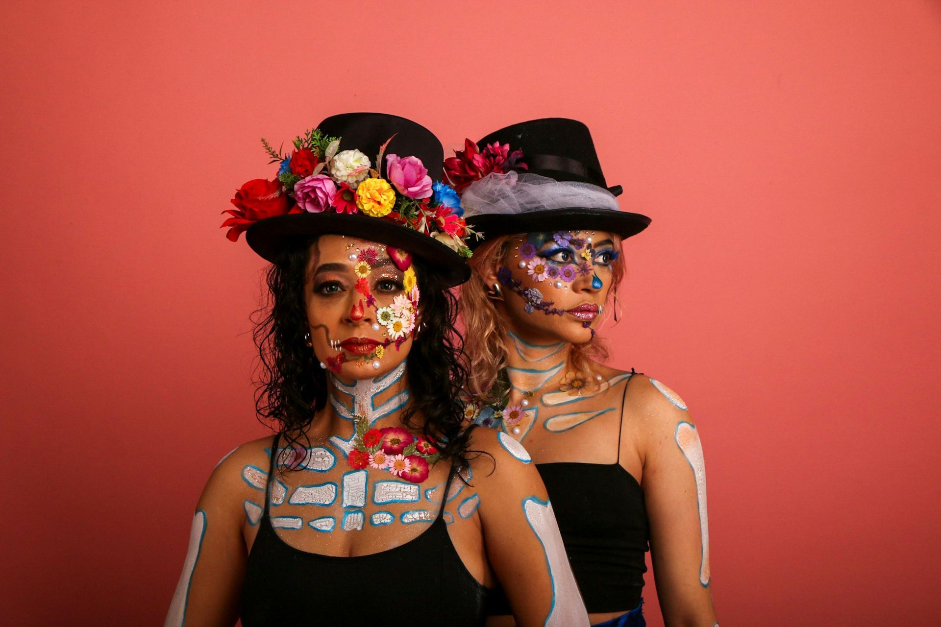 Twee mensen met Día de Muertos-make-up en -hoeden poseren voor een roze achtergrond.