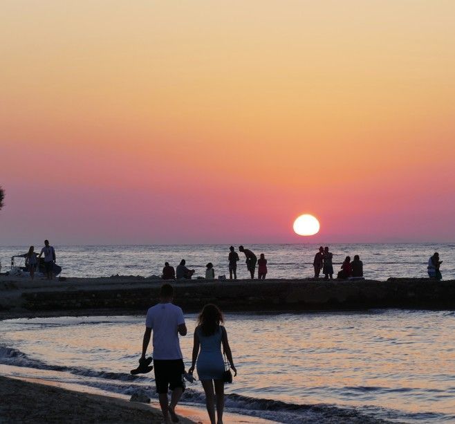 Een stel loopt over het strand bij zonsondergang; andere mensen staan ​​op de pier; een oranje en roze lucht.