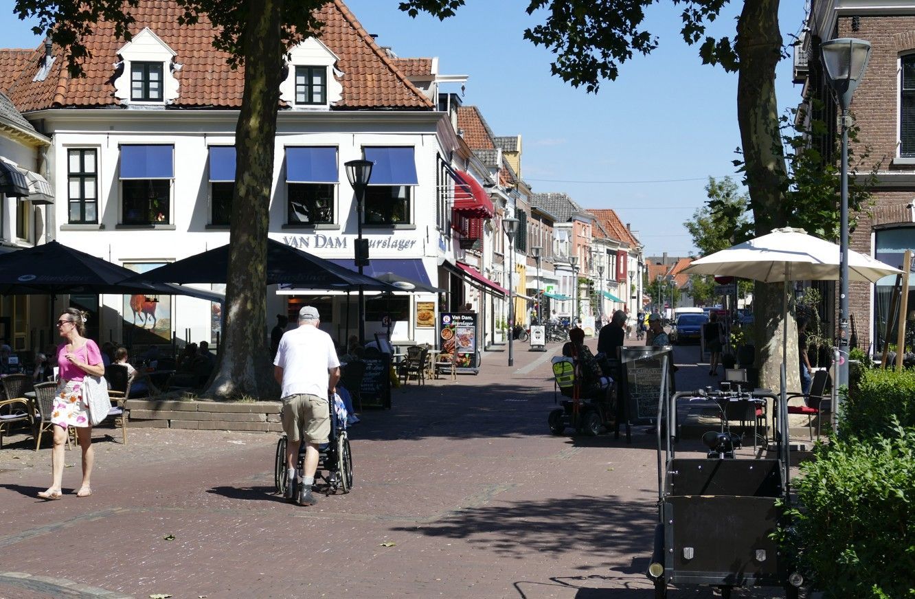 Mensen lopen over een bakstenen straat met winkels en terrasjes in een Europees stadje. Blauwe luifels, zonnige dag.