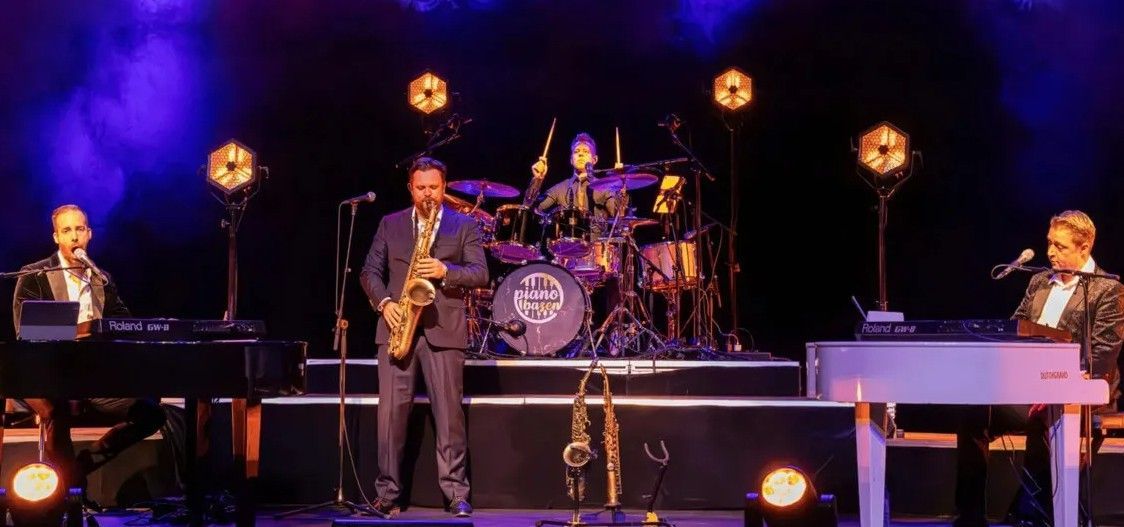 Een band treedt op een podium op met keyboards, drums en saxofoon in een concertsetting met spotlights.