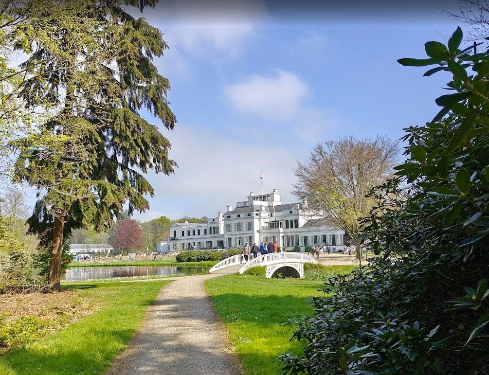 Wit herenhuis met een brug over het water. Mensen lopen over een pad in een groen park onder een blauwe hemel.