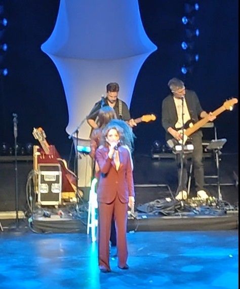 Zangeres in rood pak treedt op met band op het podium, met blauwe lichten op de achtergrond.