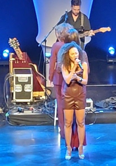Een vrouw in een bruine jurk zingt op het podium, begeleid door muzikanten.