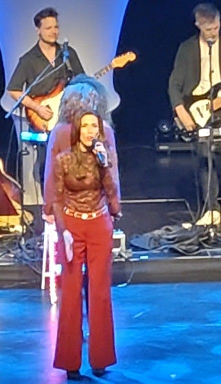 Een vrouw zingt op het podium, gekleed in een rode broek en een bruine top, terwijl op de achtergrond een band gitaar speelt.
