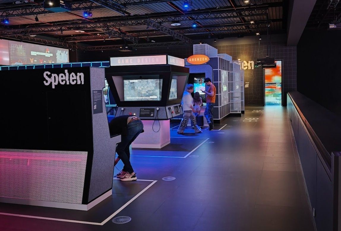 Een persoon speelt een retro arcadespel in een museumtentoonstelling. De tentoonstelling heeft ook andere speelstations.