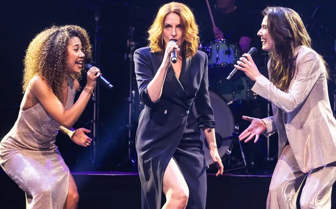 Drie vrouwen zingen op het podium met een band op de achtergrond.
