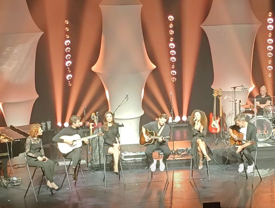 Een band treedt op het podium op. Zes mensen zitten, sommigen spelen op akoestische gitaren.