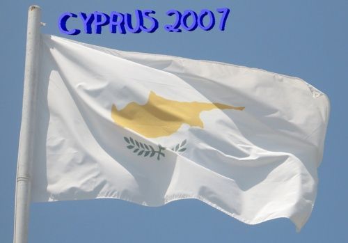 Witte Cypriotische vlag met kaart en olijftakken, wapperend aan een vlaggenmast tegen een blauwe lucht.