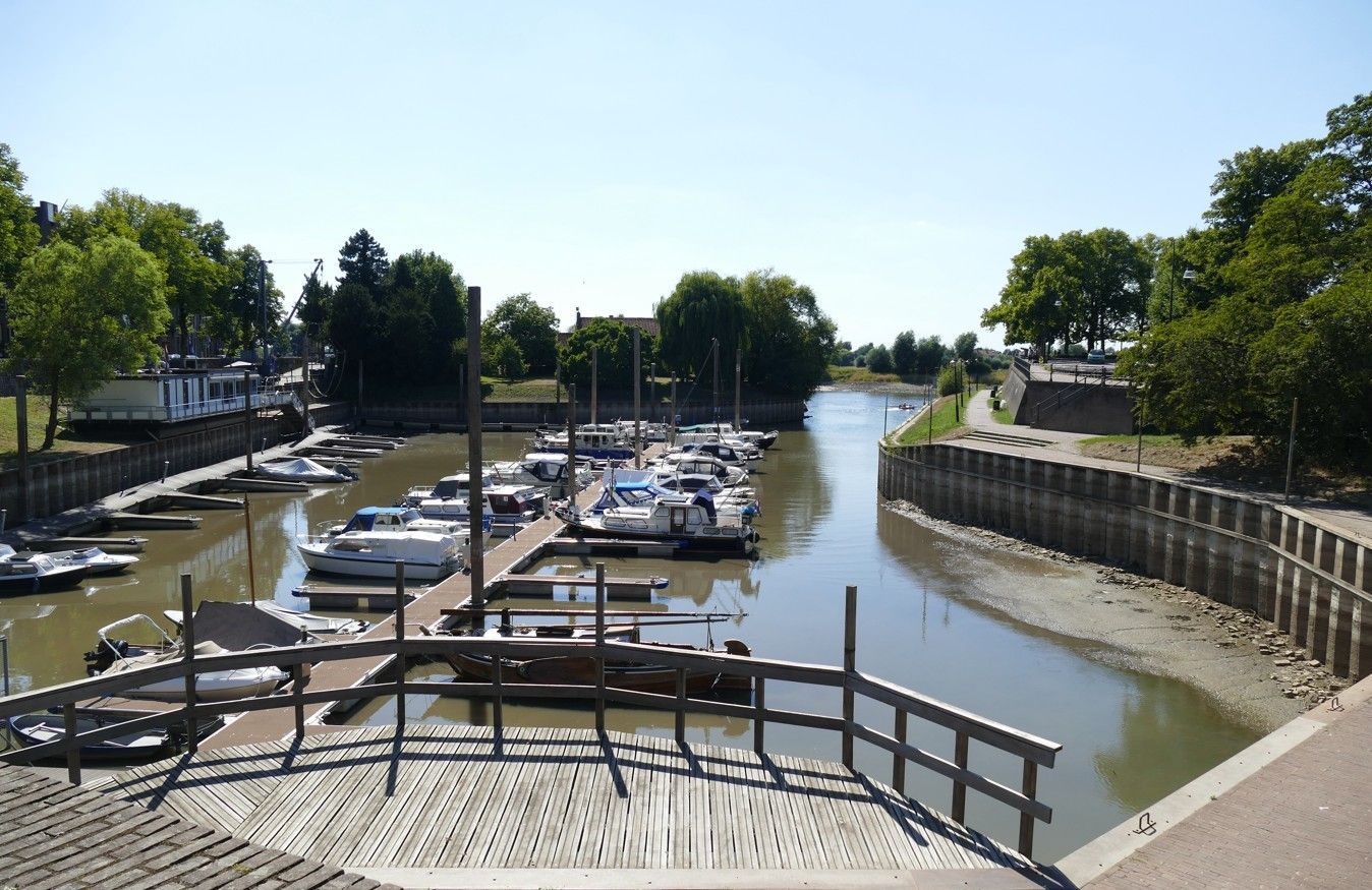 Boten liggen aangemeerd in een waterweg, omzoomd door steigers en bomen, onder een helderblauwe hemel.
