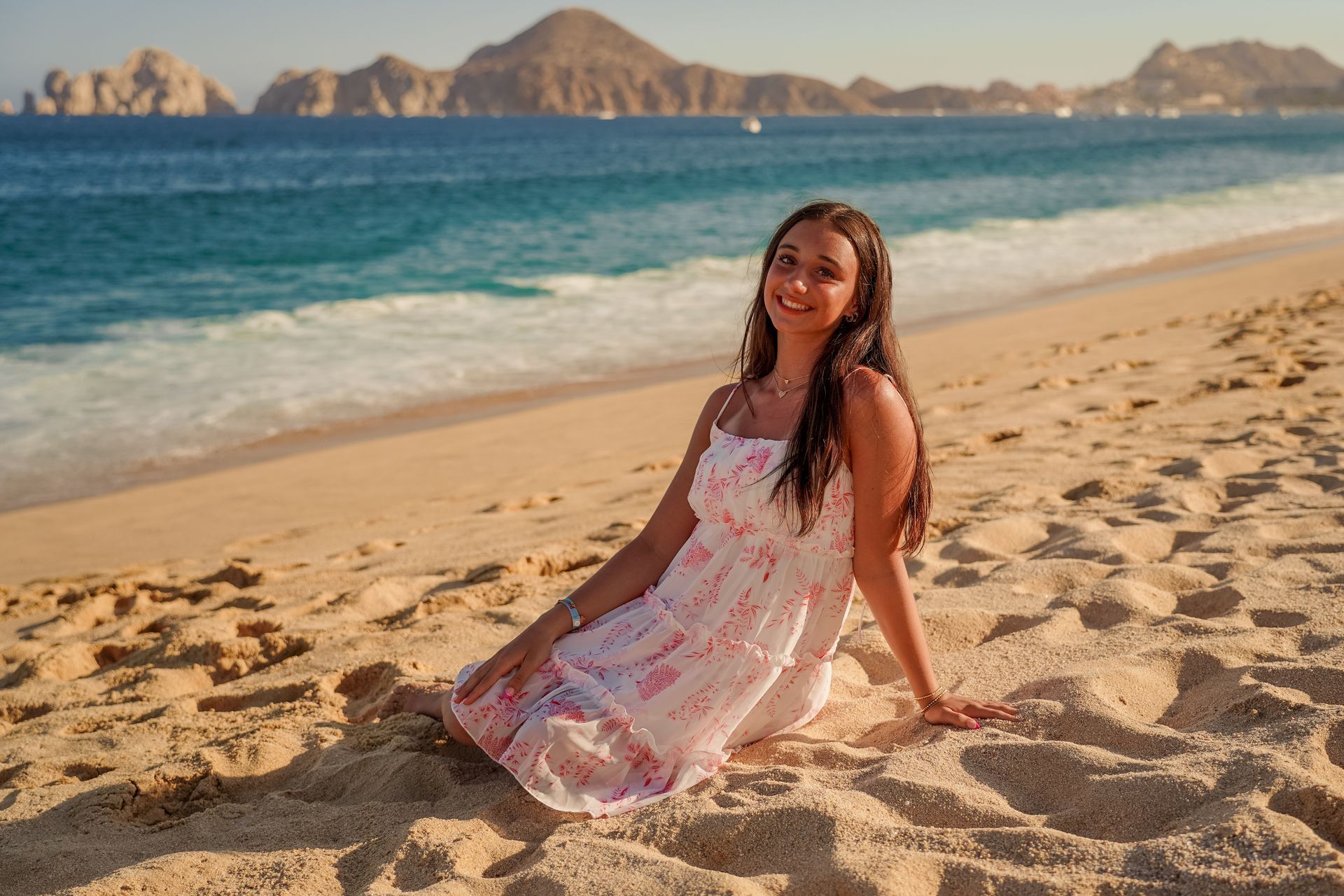 Woman in a white dress smiles on a sandy beach; turquoise water and mountains in the background.