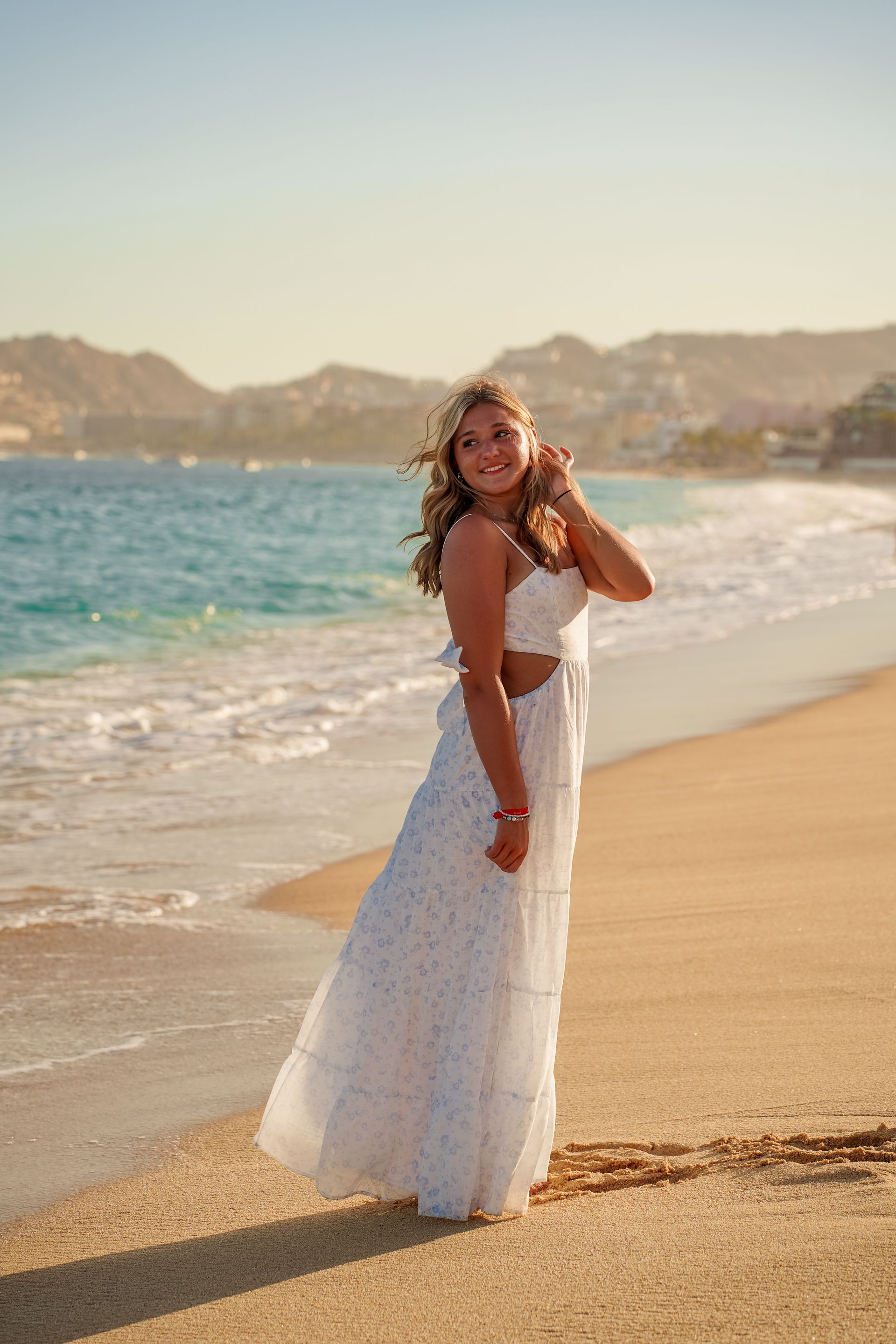 Woman in white dress on beach, smiling. Ocean and mountains in background.