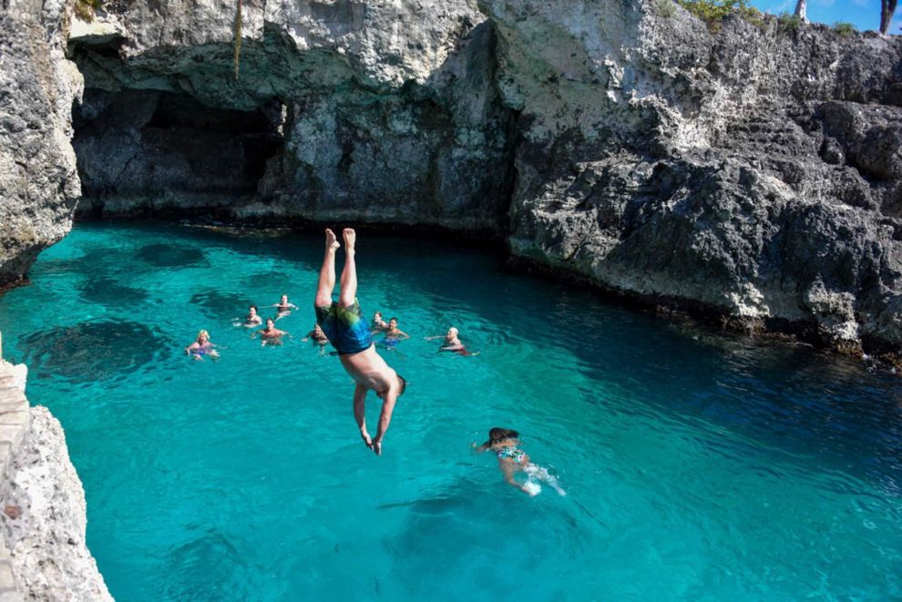 Man doing a backflip into turquoise water, other people swimming nearby, cliffside setting.