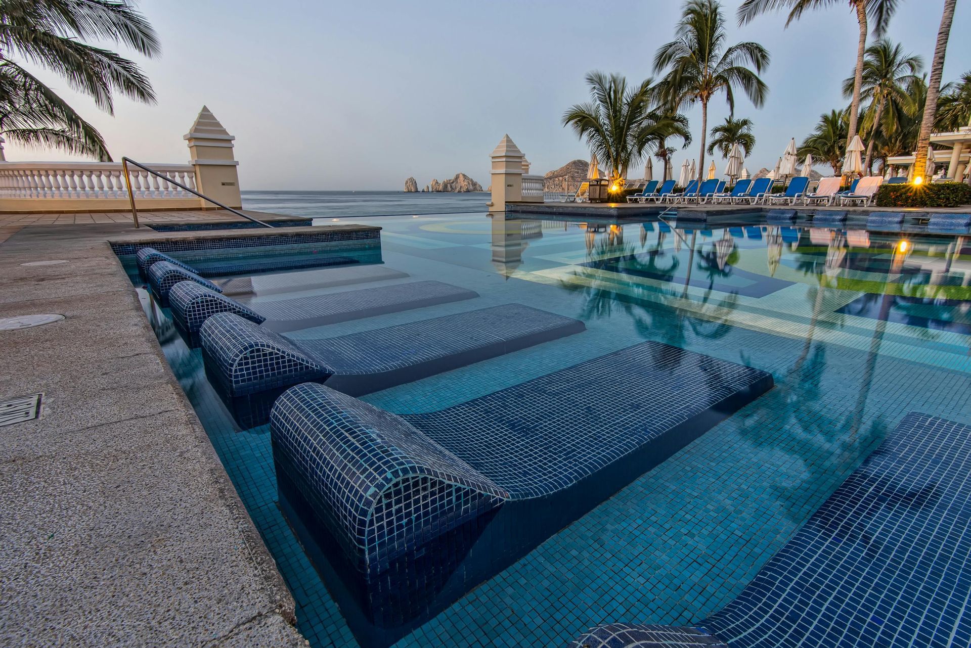 Pool with blue tiled loungers, ocean view, palm trees.