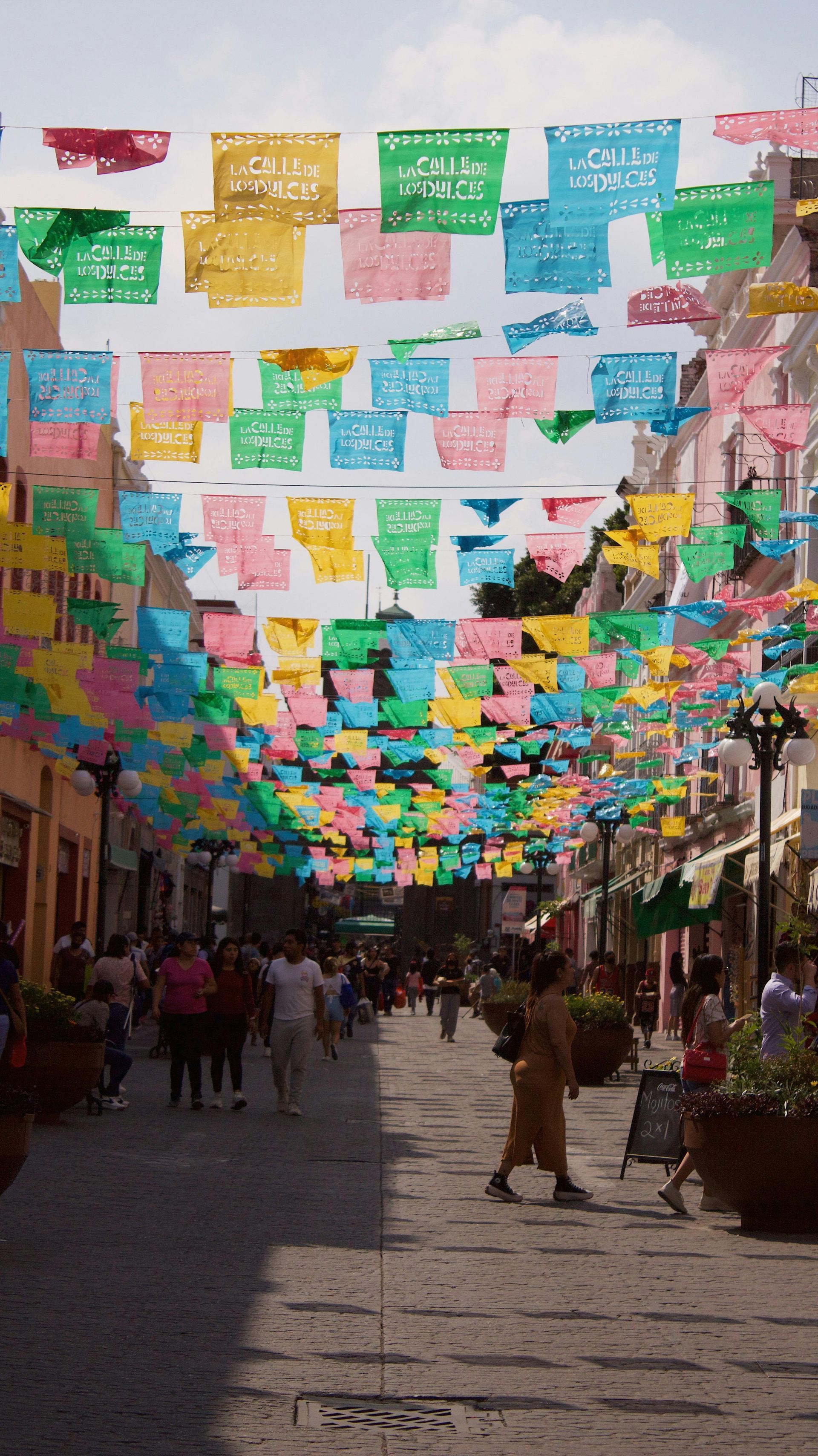Colorful papel picado decorations line a pedestrian street in Mexico, people stroll beneath the banners.