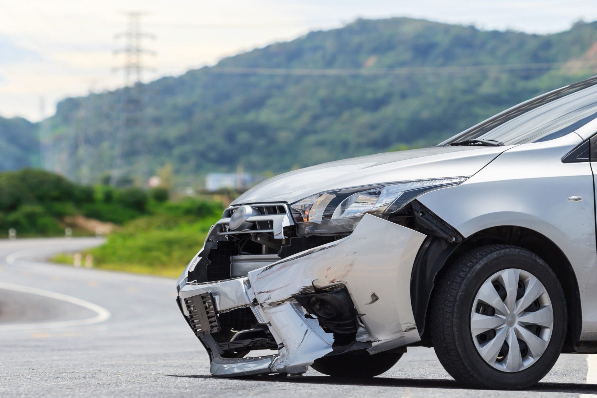 A silver car with a damaged front bumper is parked on the side of the road.