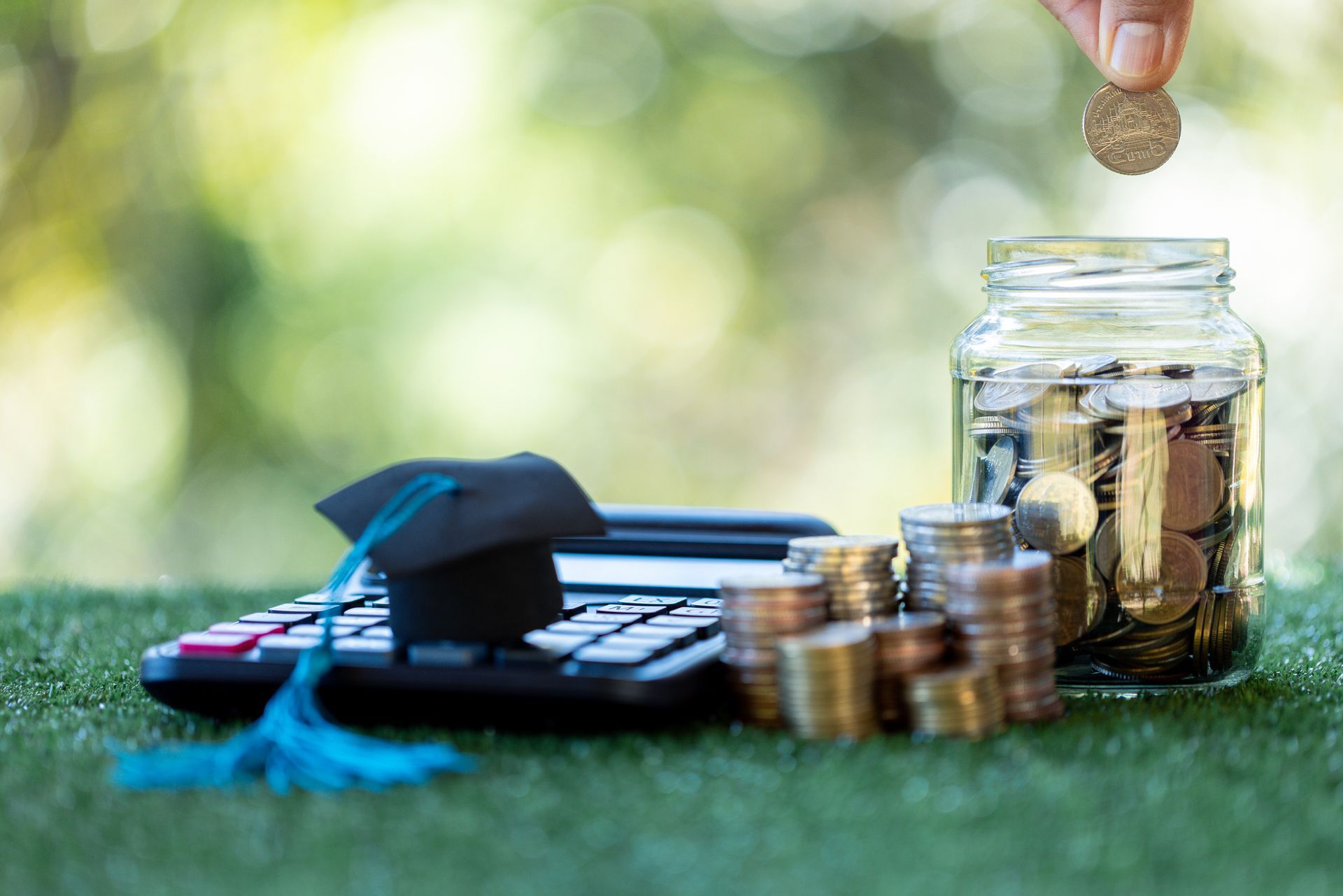 Hand placing a coin into a jar beside a calculator and stacks of coins on grass