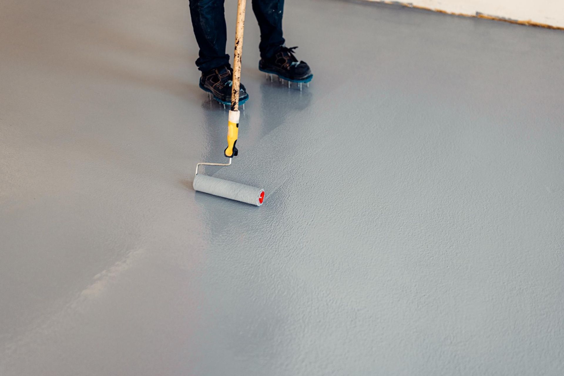 Person using a paint roller on a gray floor, applying white coating near their boots