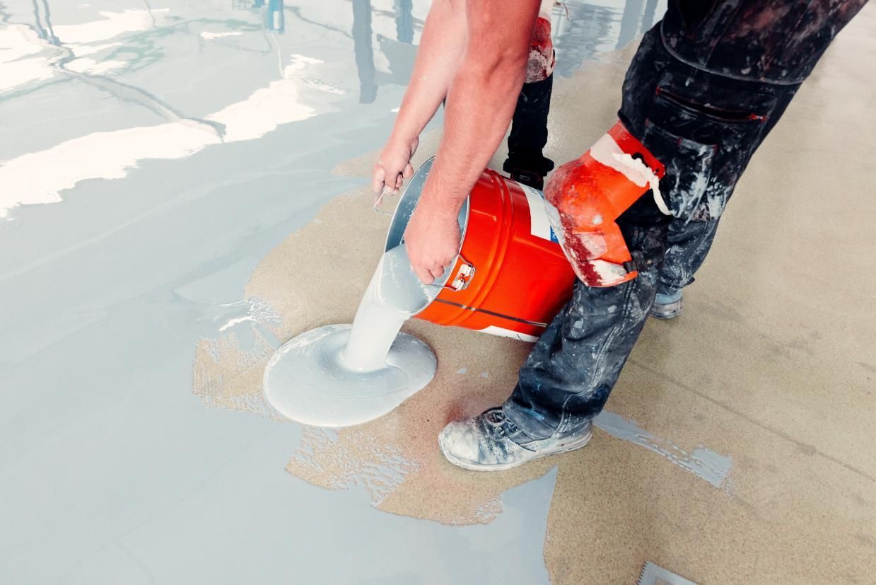 Pouring gray floor leveling compound from an orange mixer onto a concrete floor