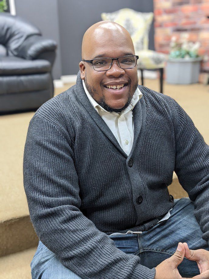 Man wearing glasses and gray sweater, smiling, sitting on steps indoors.