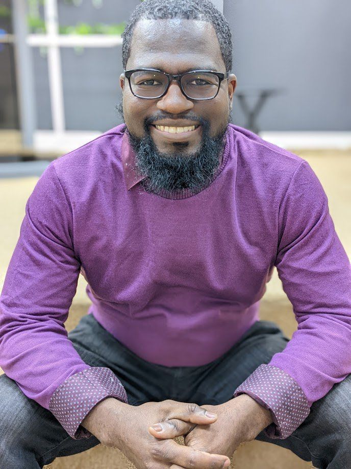 Man in purple shirt and glasses smiles, sitting outdoors.