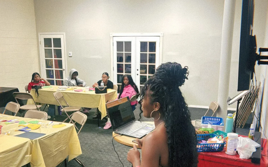 Woman speaking with laptop at a table, with a group of people at tables in a room with light-colored walls and doors.