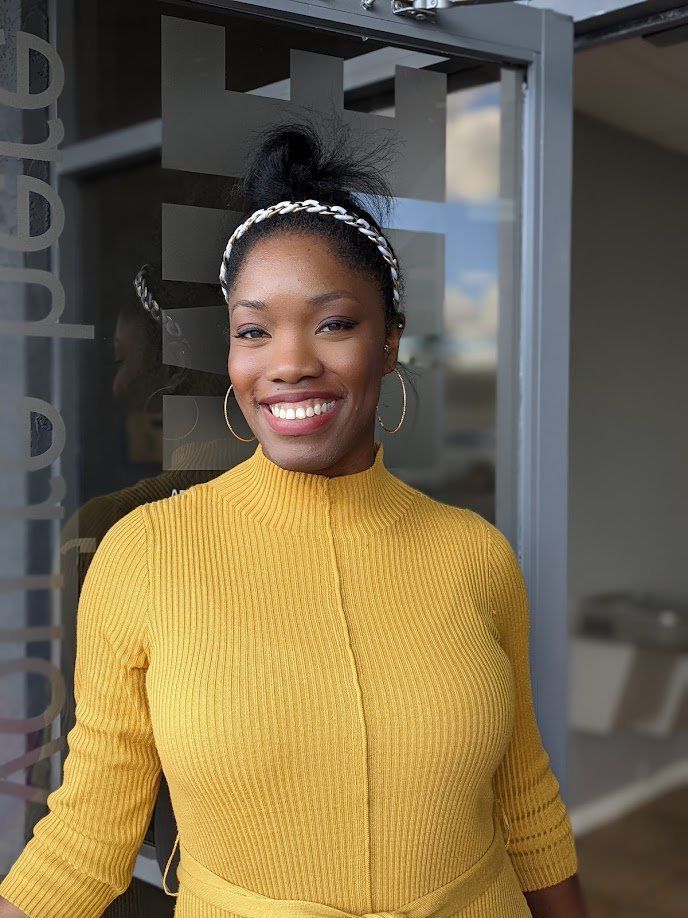 Woman with dark hair in yellow dress, smiling, wearing headband and hoop earrings. Standing near glass door.