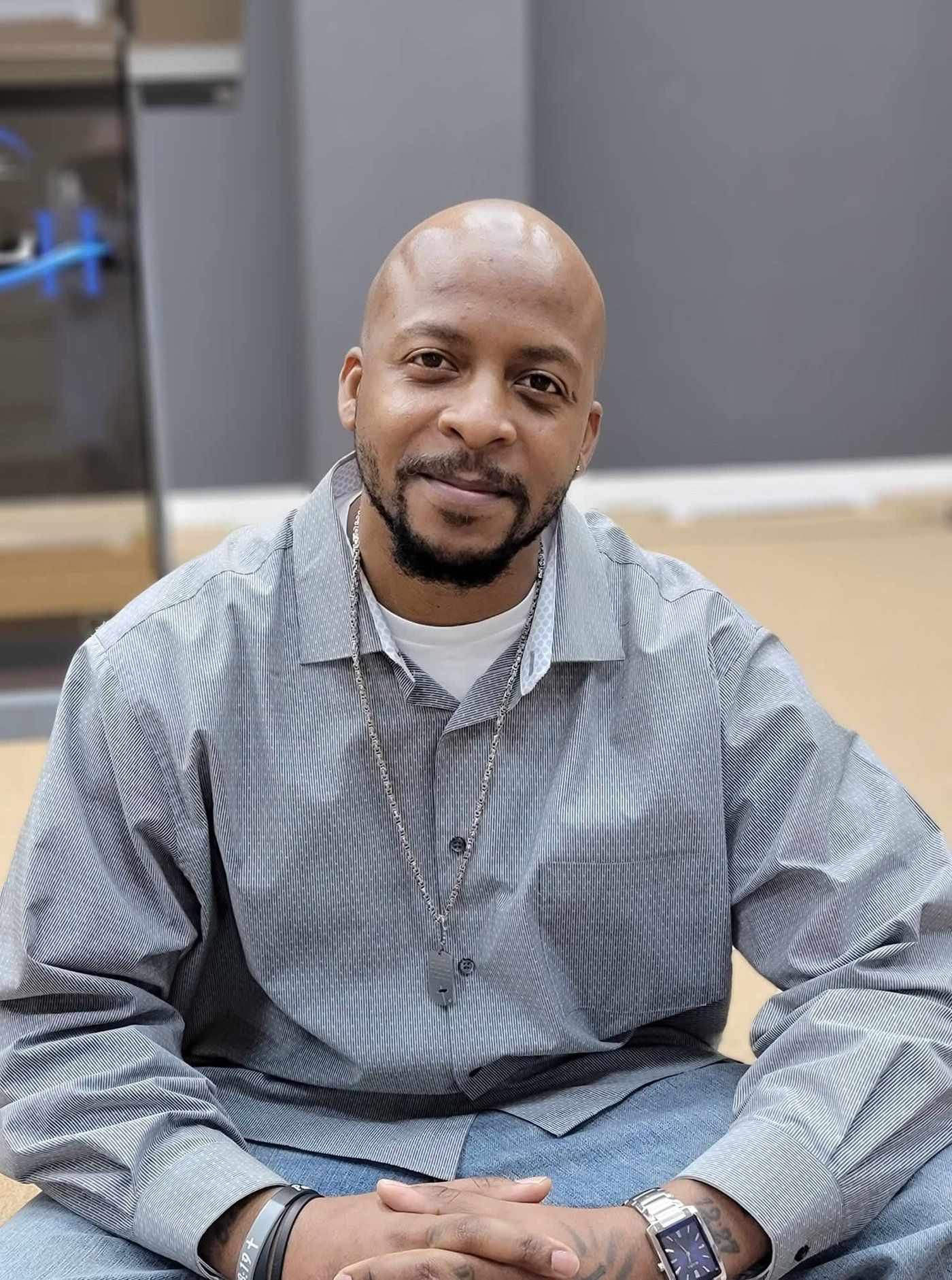 Man with shaved head, smiling, in striped button-up shirt and jeans, sitting.