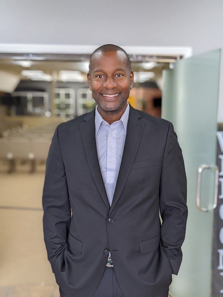 Man in a suit smiles, hands in pockets. Standing near glass doors, modern interior.