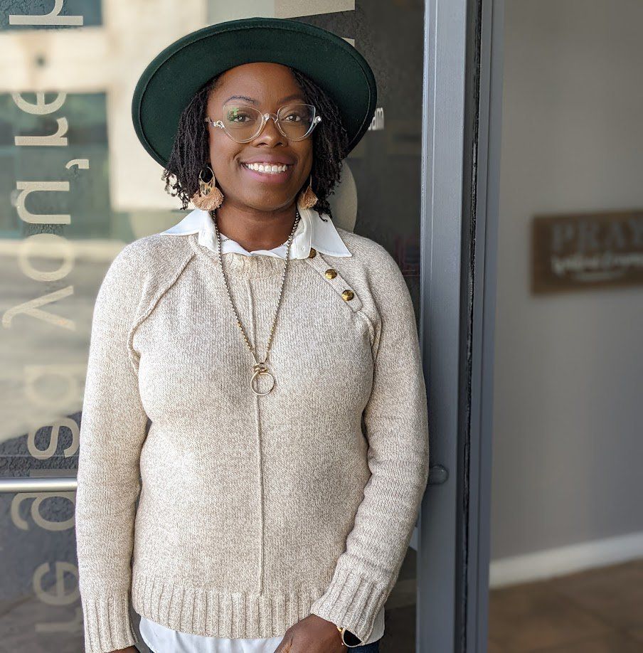 Woman in a green hat and glasses smiles, leaning against a door. She wears a beige sweater and gold necklace.