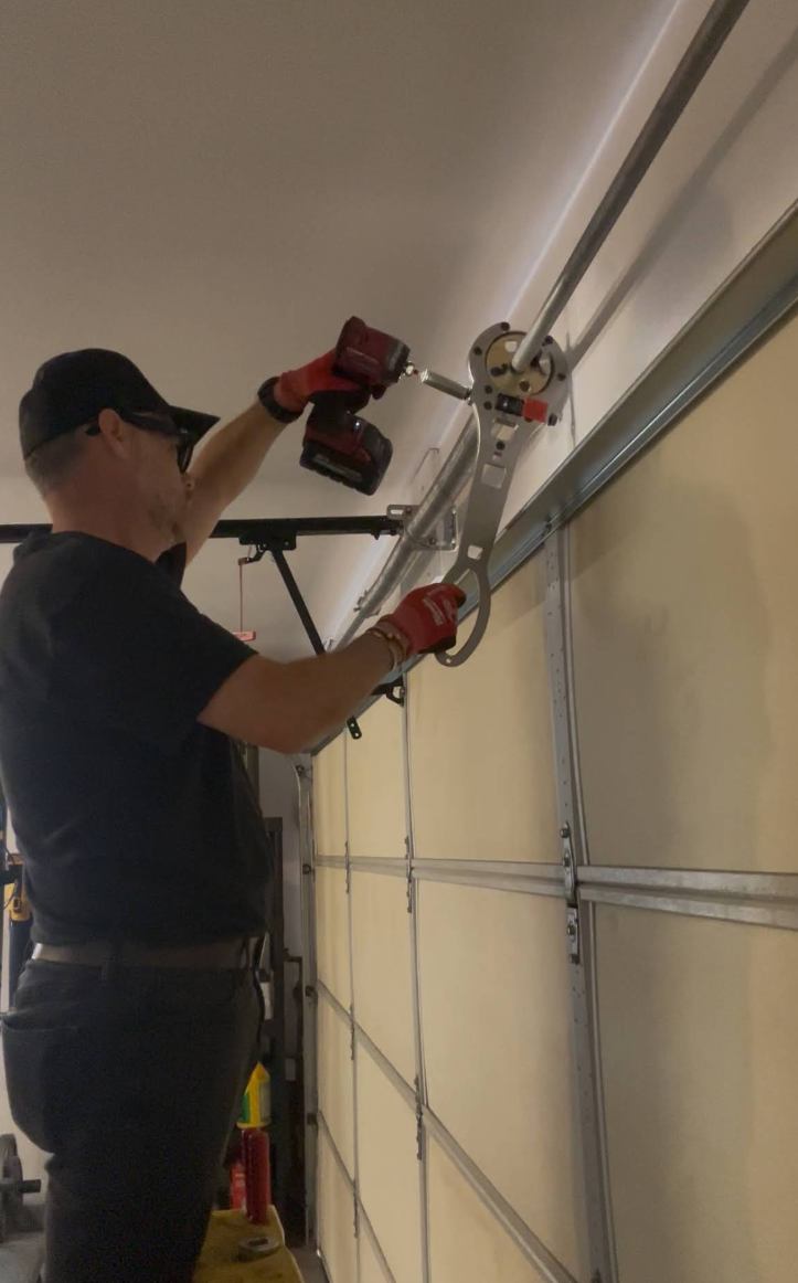 Man installing a garage door opener on a ladder, using a red power drill.