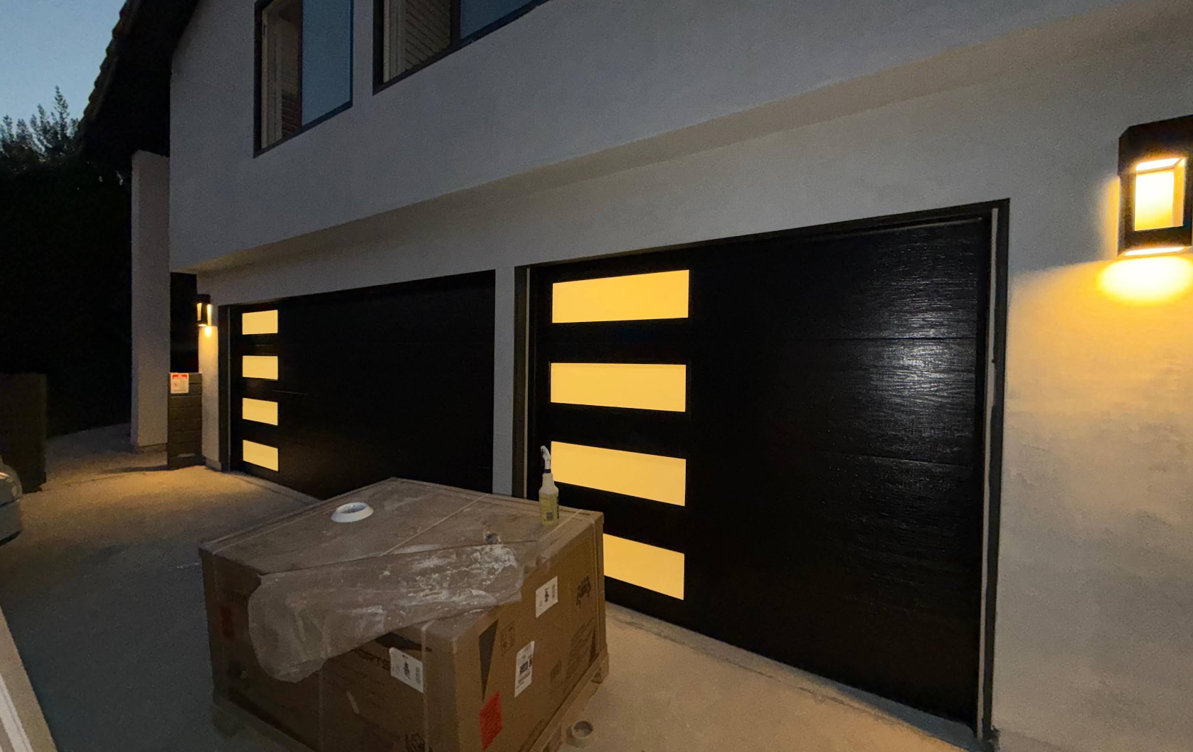 Nighttime view of a building facade with black garage doors, lit windows, and a cardboard box on the ground.