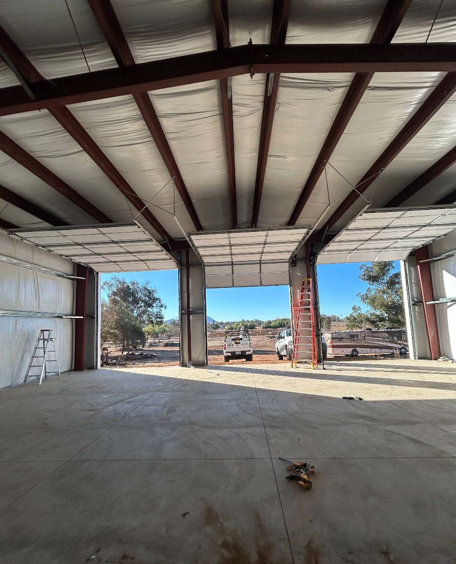 Empty steel-framed warehouse under construction with open doors and concrete floor, sunlight outside