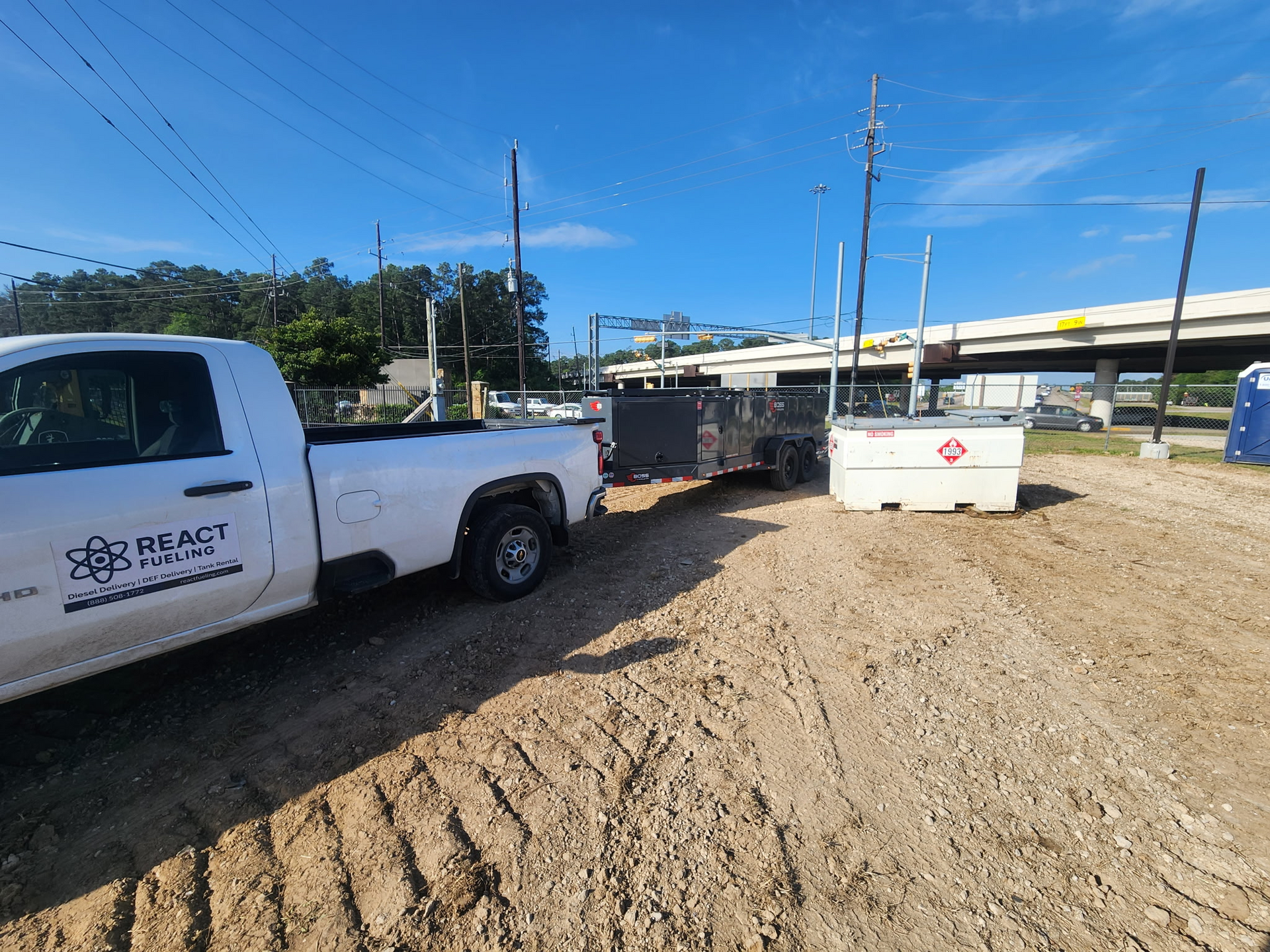 White pickup and utility trailer on a dirt lot beside a road under a clear blue sky.