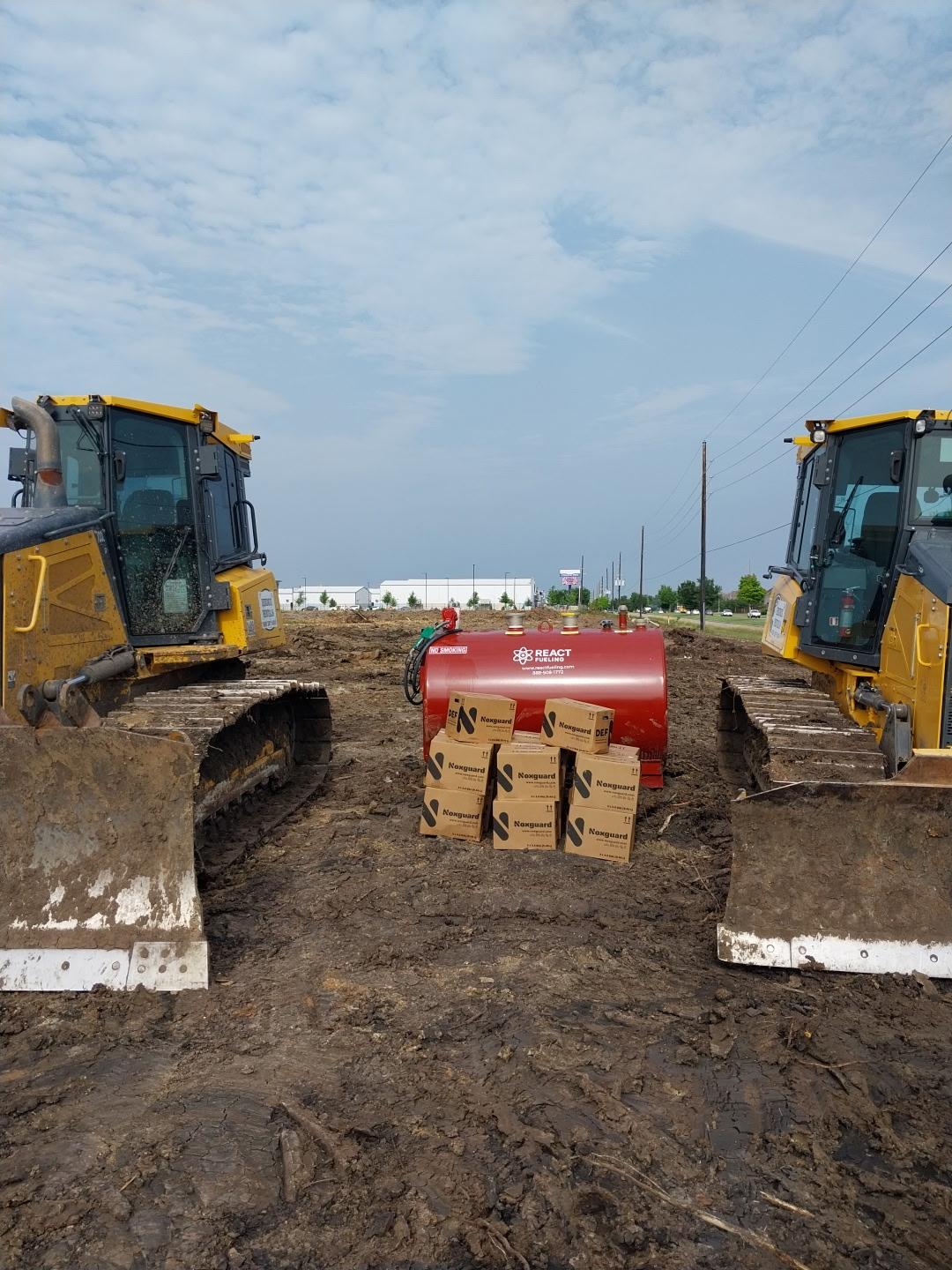 Two yellow bulldozers face a red farm implement in a muddy field under a cloudy sky.