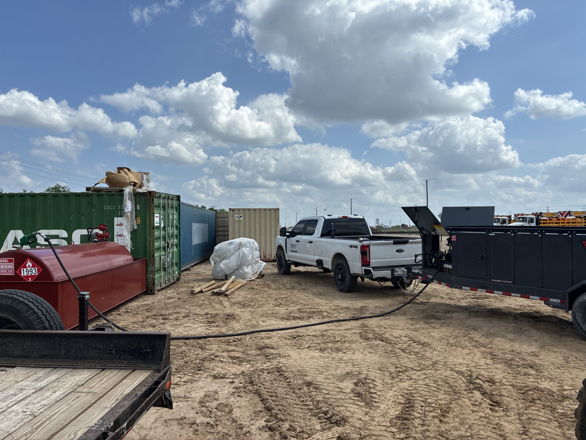 White pickup and red truck beside dumpsters in a muddy lot under a cloudy sky