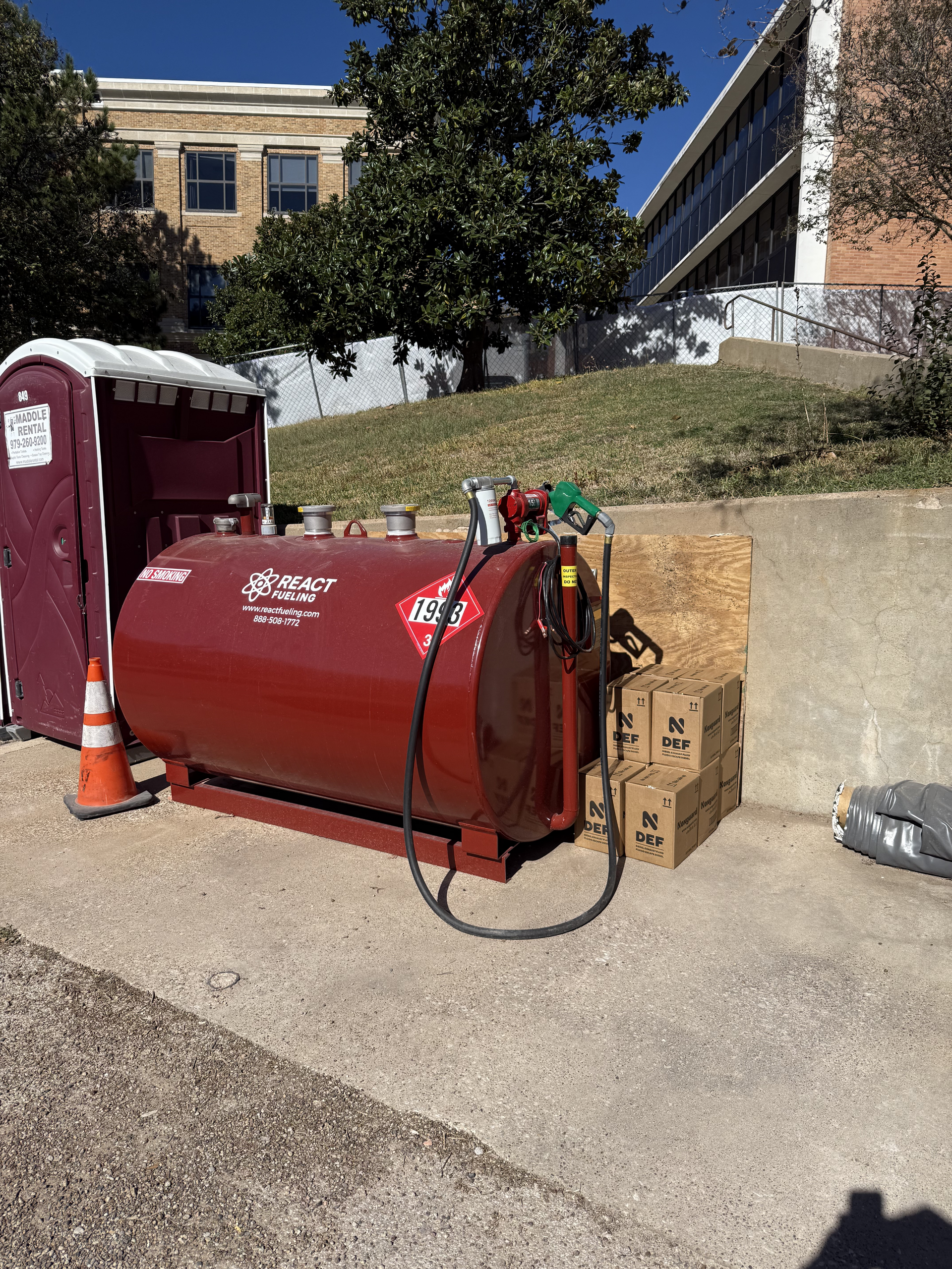 Red cylindrical tank with hoses and boxes on a sidewalk beside a building and tree.