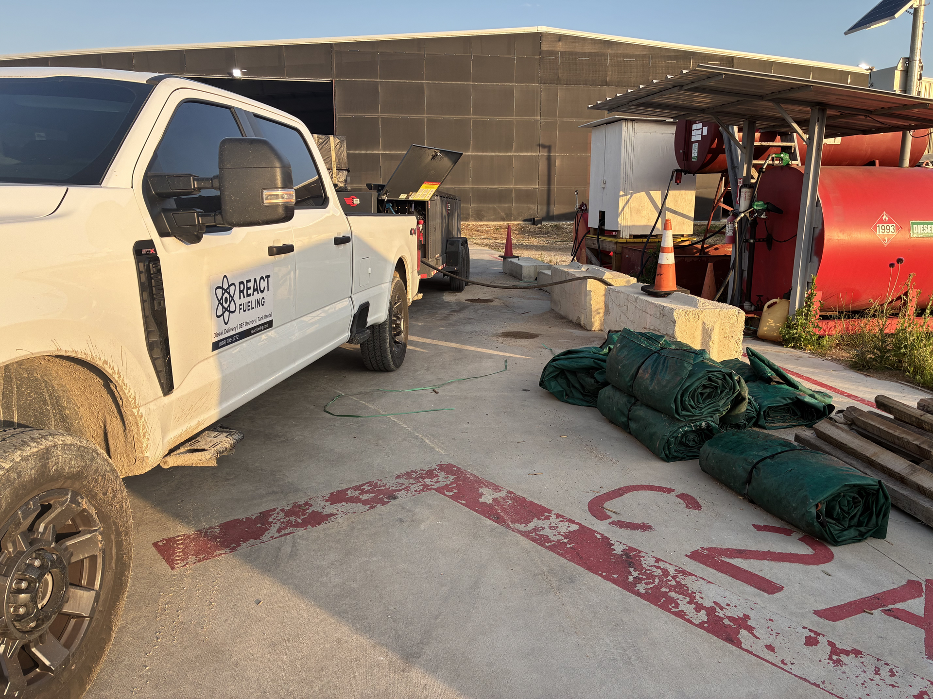 White pickup truck beside red equipment and green rolled tarps on a concrete lot with a wall and canopy in back