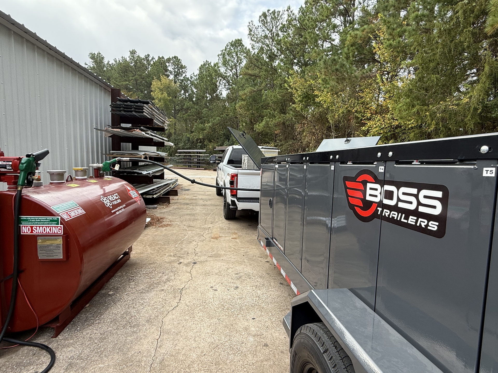 White pickup and black BOSS trailer in a gravel lot beside stacked materials and a red tank, near a metal building