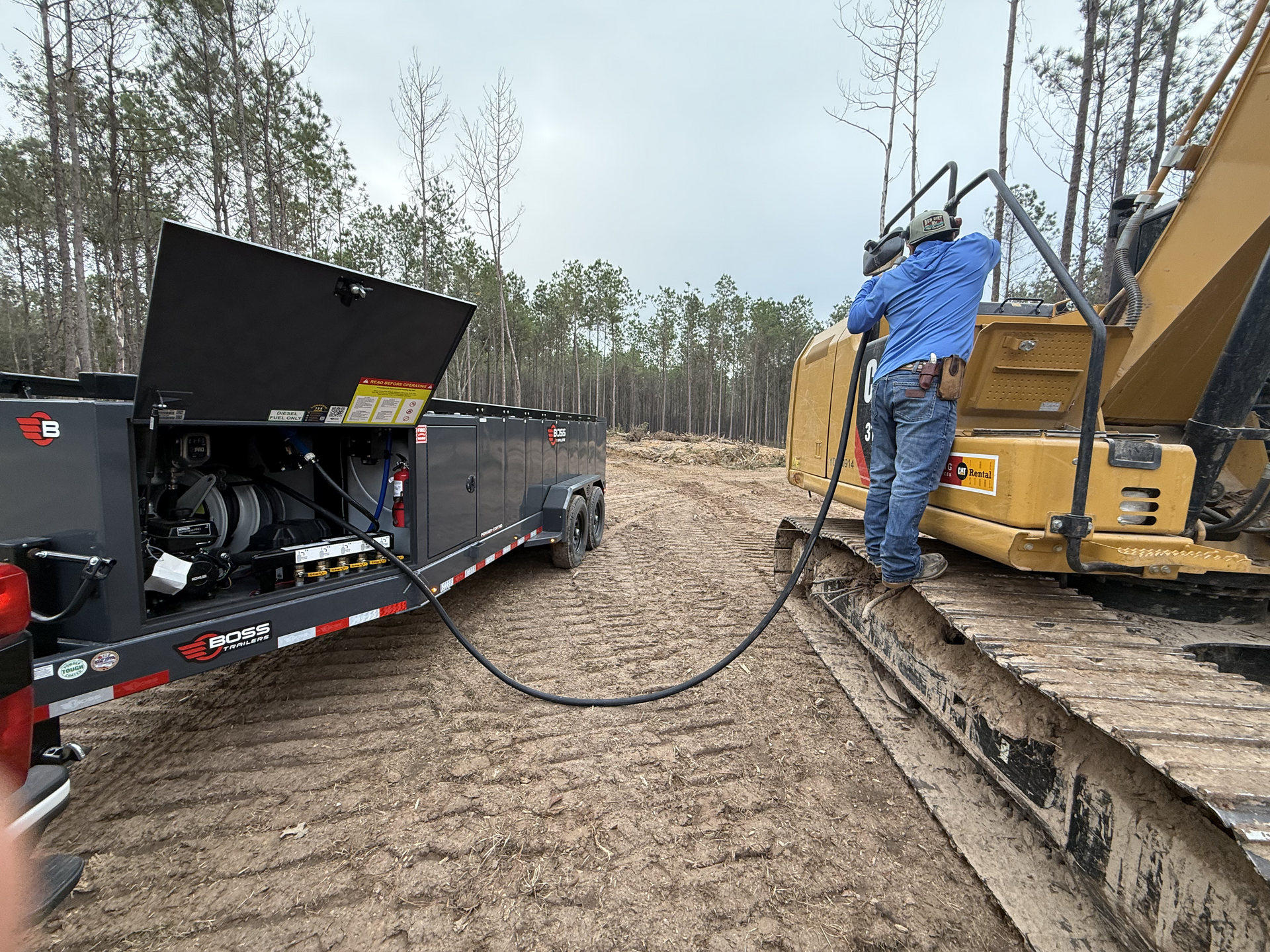 Heavy equipment and worker on muddy construction site, connected by a hose to a trailer-mounted machine