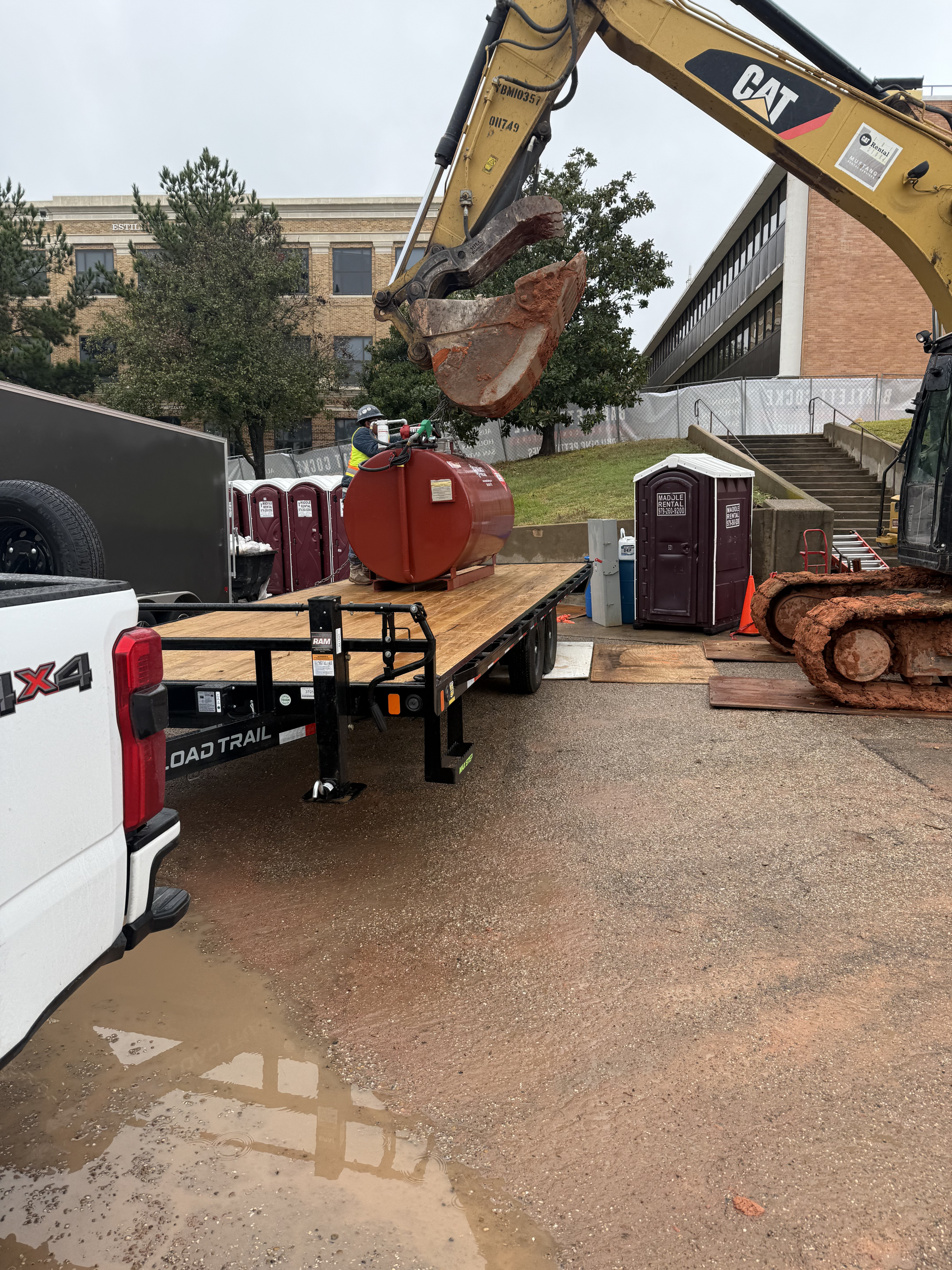 Excavator lifting a large rusted cylinder onto a flatbed trailer in a gravel lot