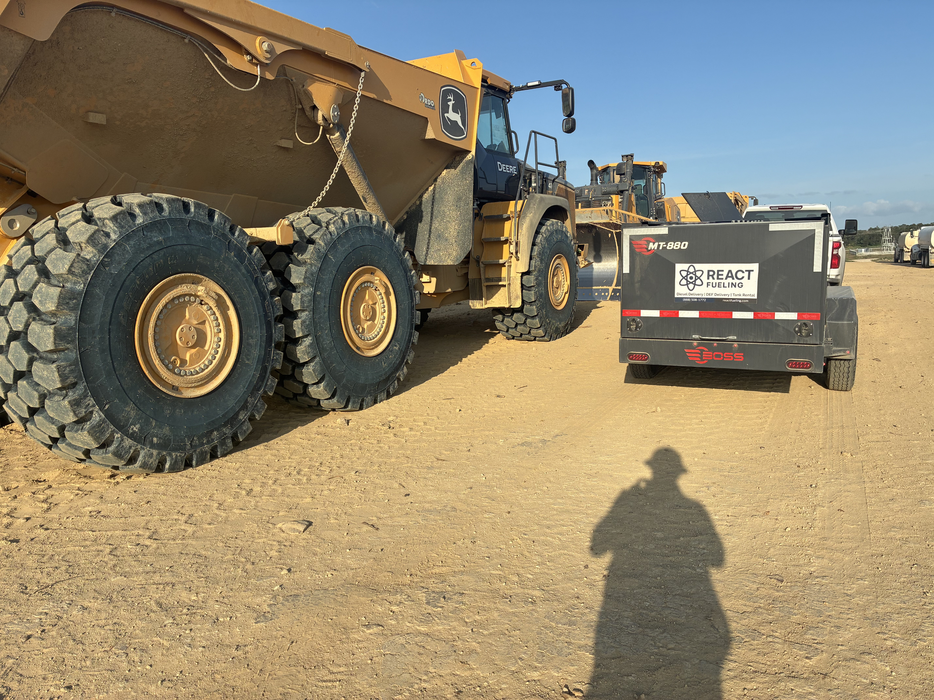 Large yellow dump truck beside a black utility trailer on a dirt construction site.