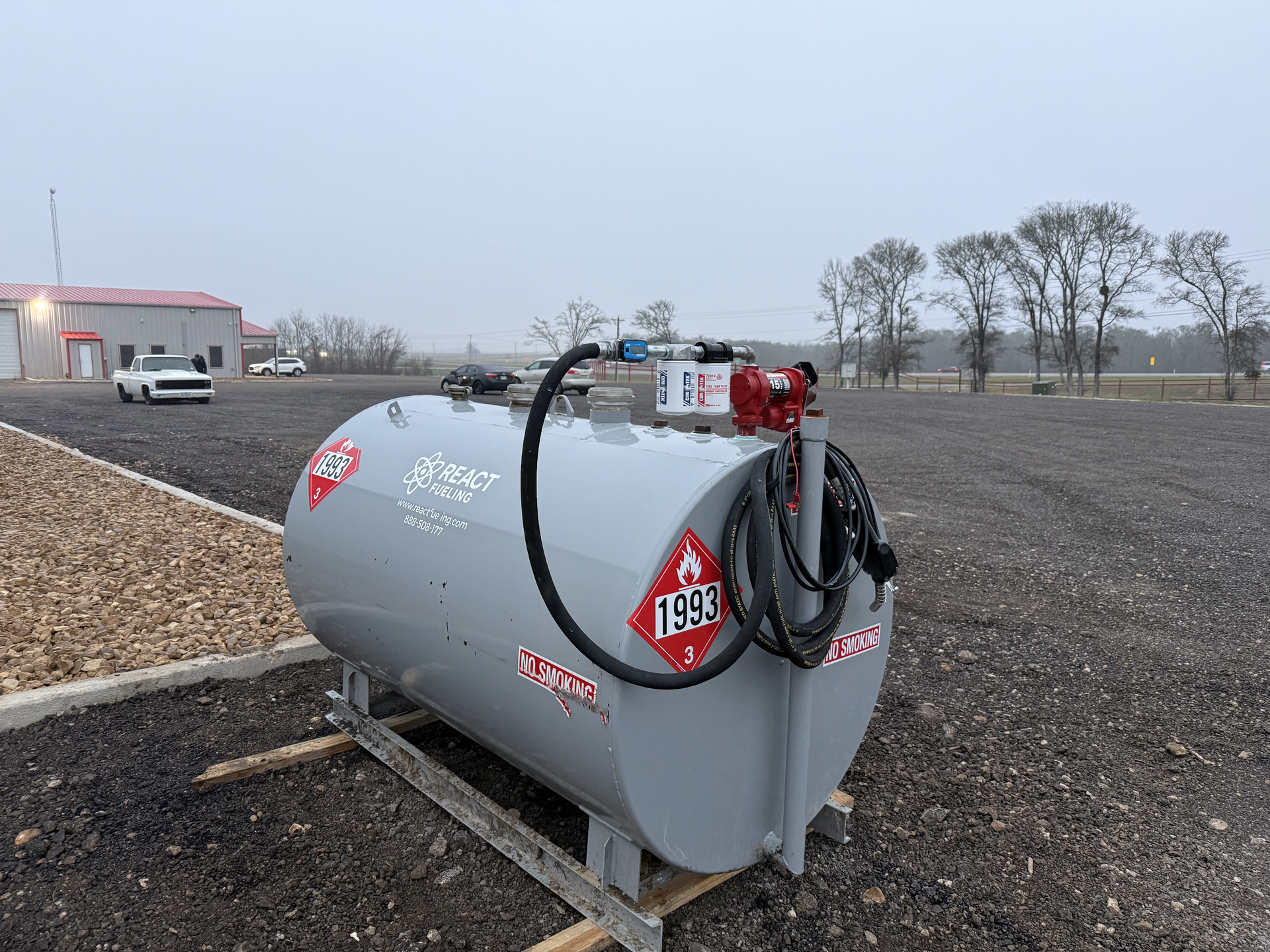 Gray fuel tanker in a gravel lot near a roadside building, with hoses and red fittings attached.
