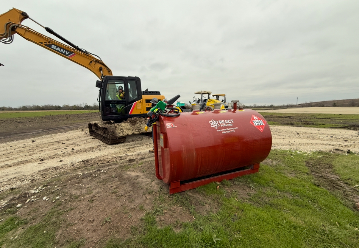 Yellow excavator beside a large red cylindrical tank in a muddy field under overcast sky
