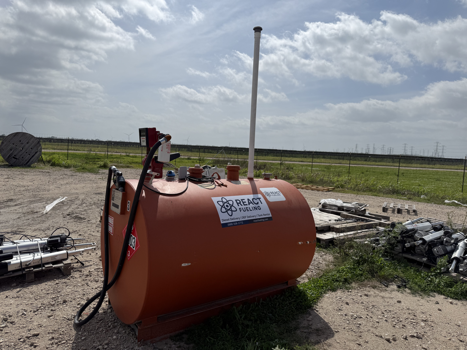 Orange cylindrical tank on a gravel field under cloudy sky, with a white pole and scattered equipment nearby