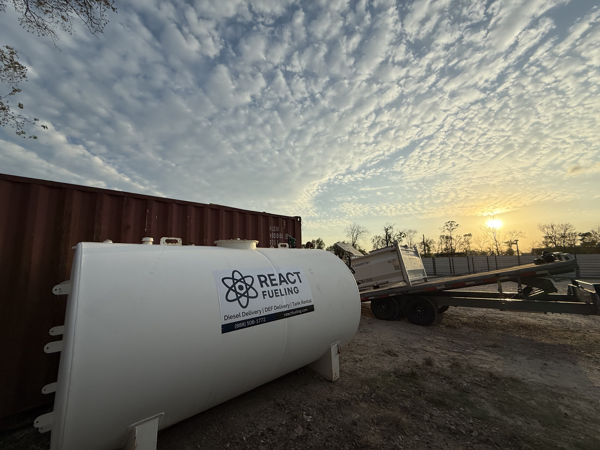 White water tank in a field at sunset with a cloudy sky and industrial equipment in the distance