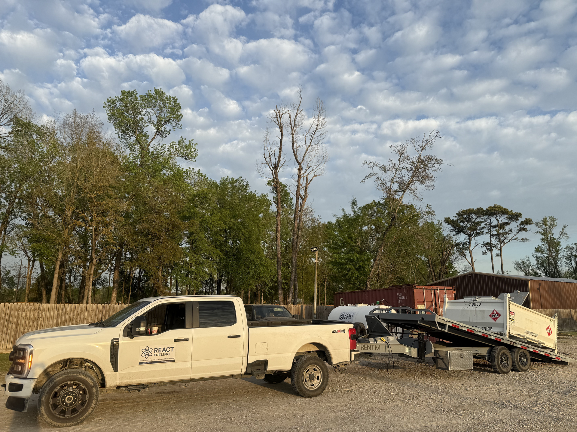 White pickup towing a trailer with a large sign, parked on a dirt lot under cloudy skies